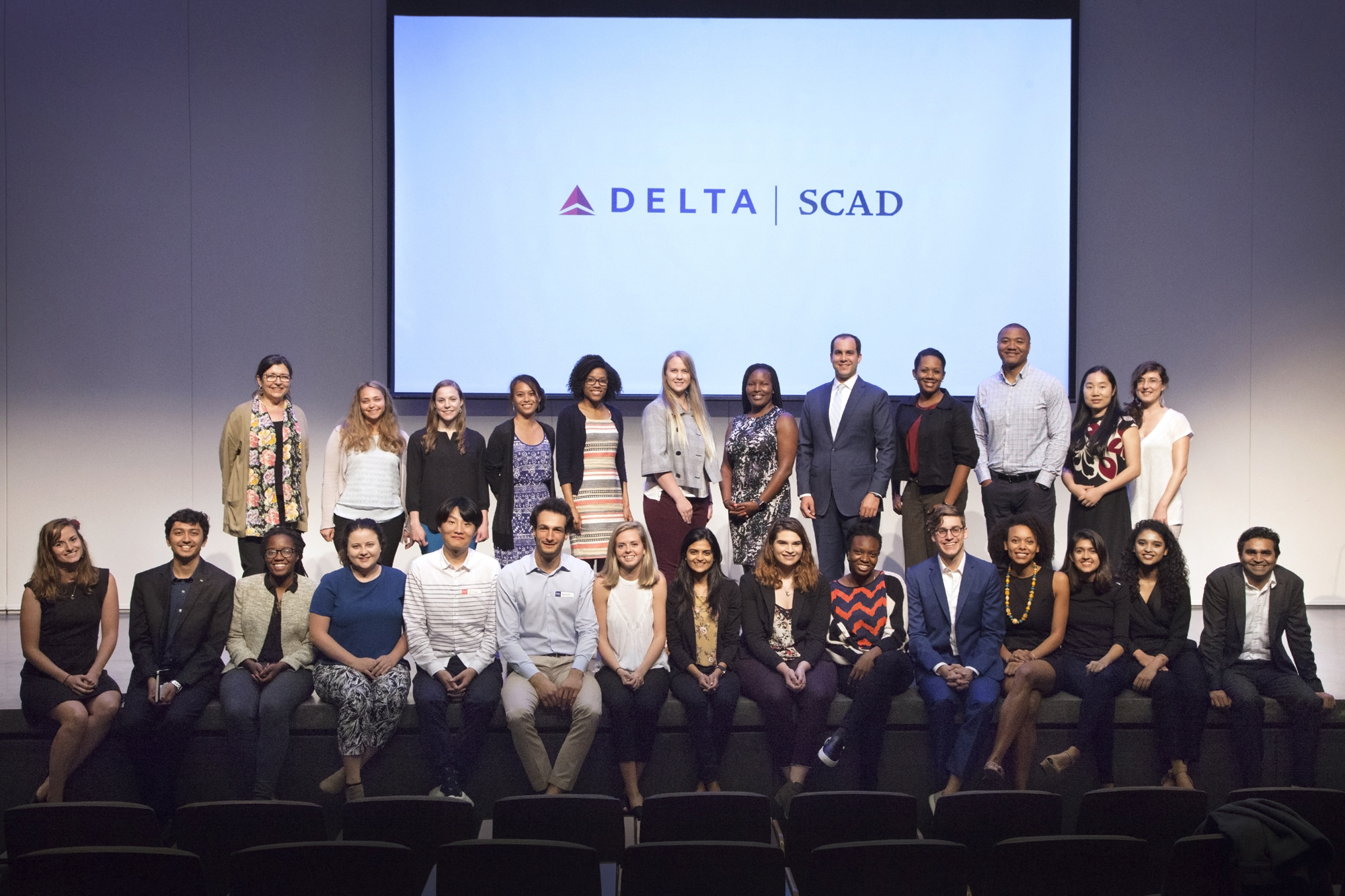 Large group of students gather onstage and pose in front of screen with the Delta and SCAD logos