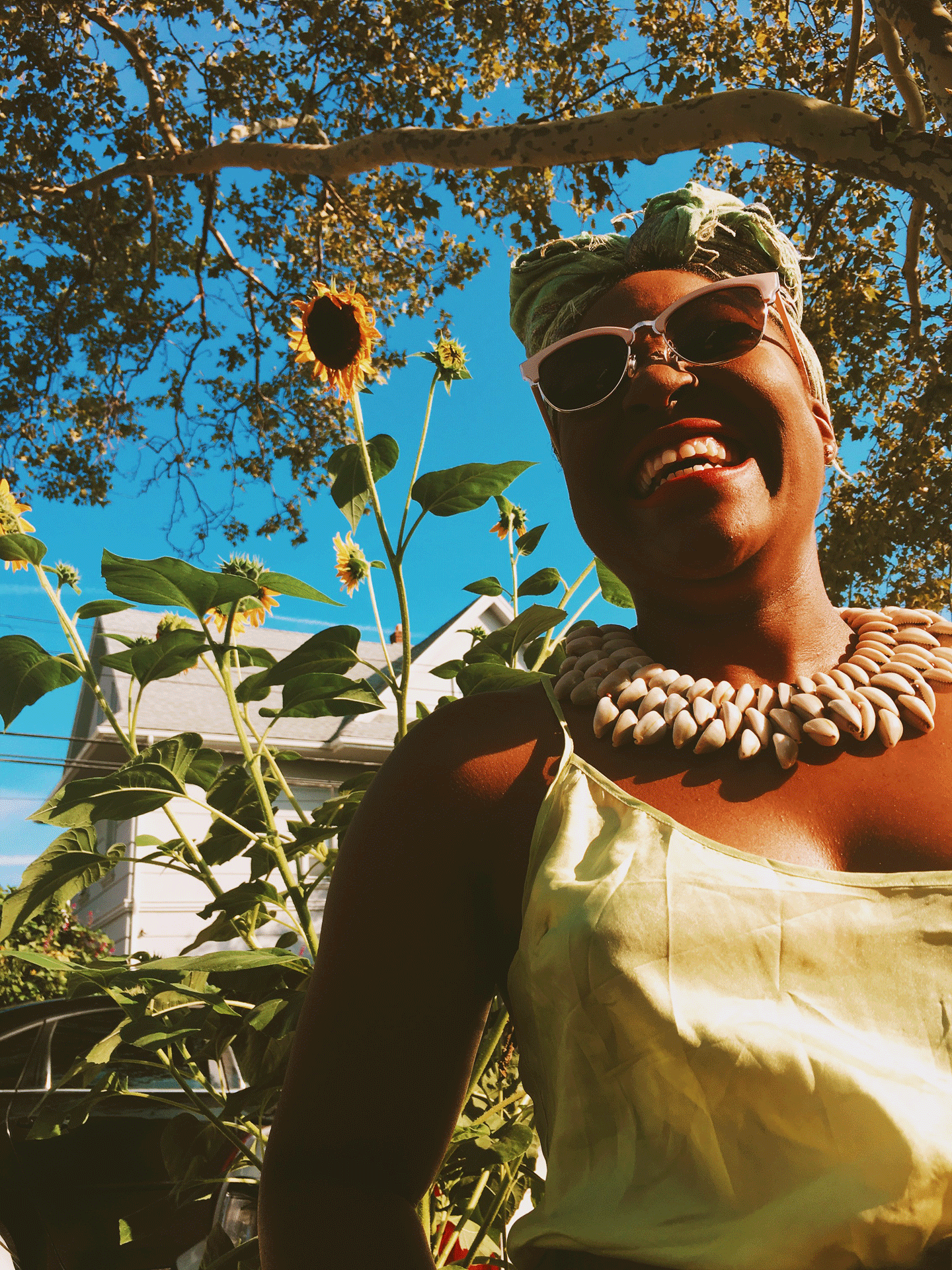 Erika Wade wears sunglasses and smiles big with tall sunflowers behind her