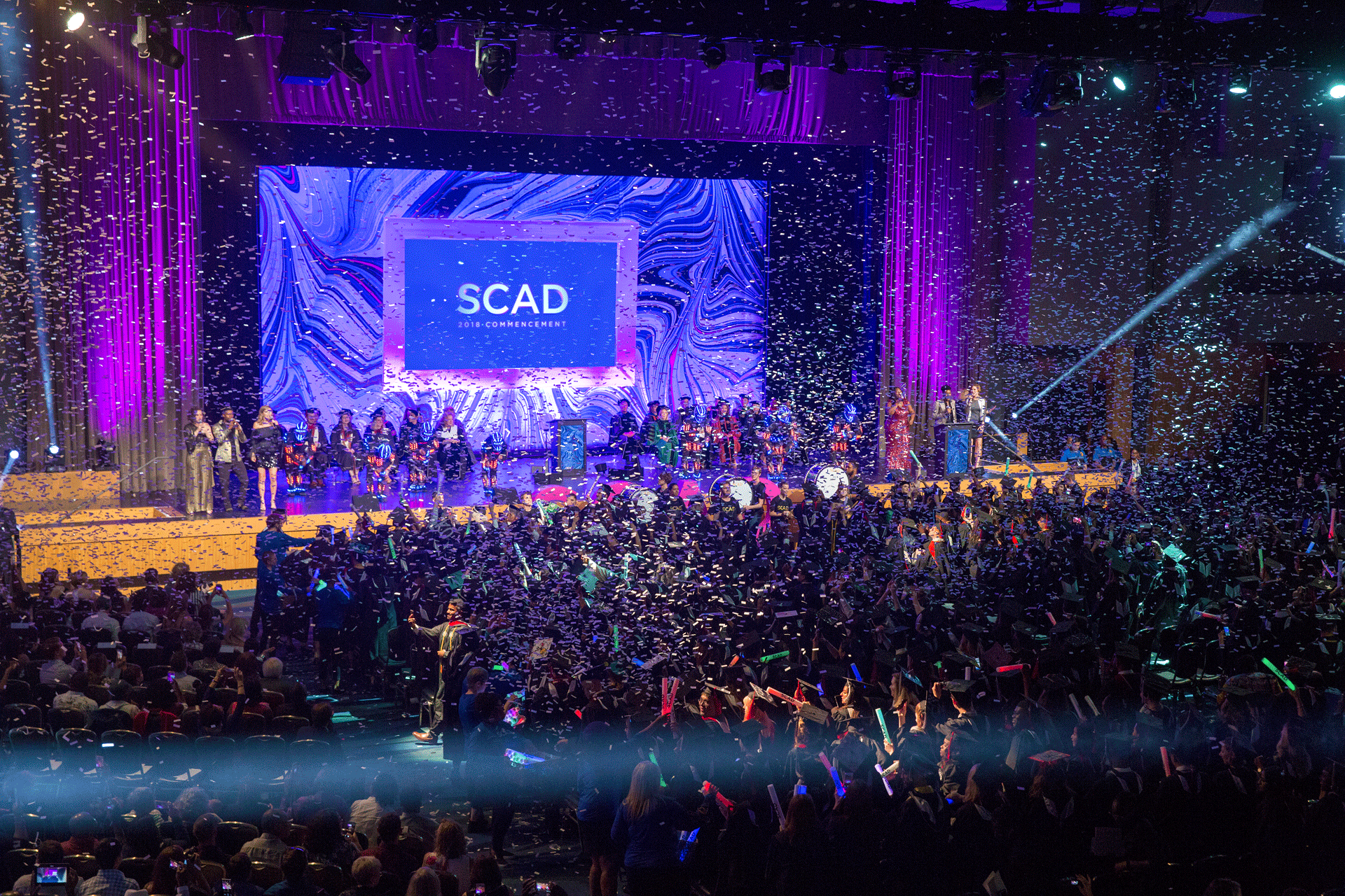 Rows of graduates celebrate with confetti in the air and SCAD banner ahead