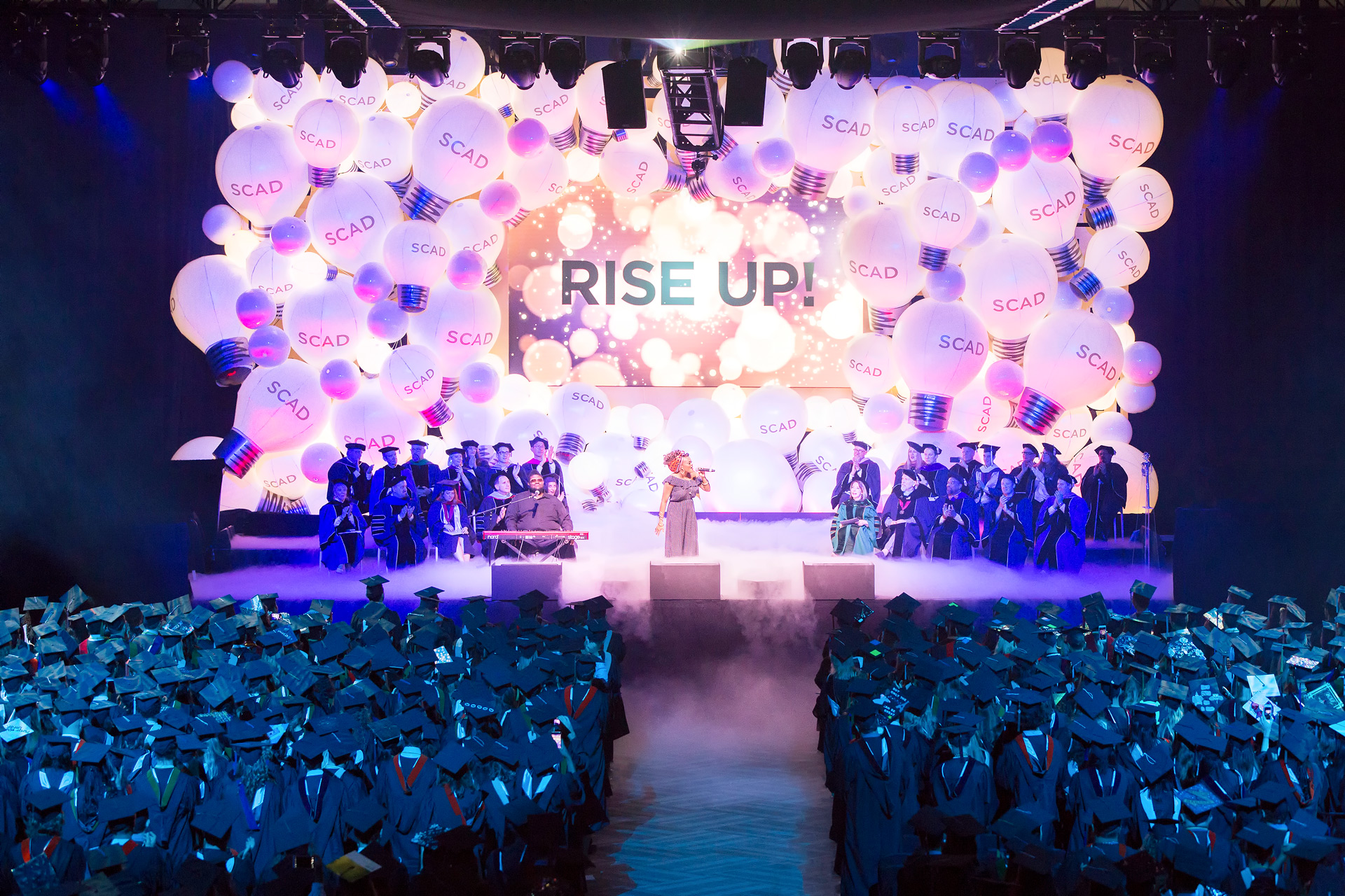 Woman sings center stage surrounded by fog with decorative SCAD lightbulbs in the background and students in cap and gown in the audience
