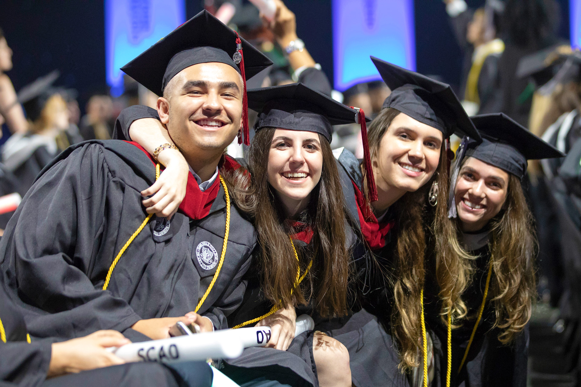 Four graduates holding diplomas and wearing cap and gown smile