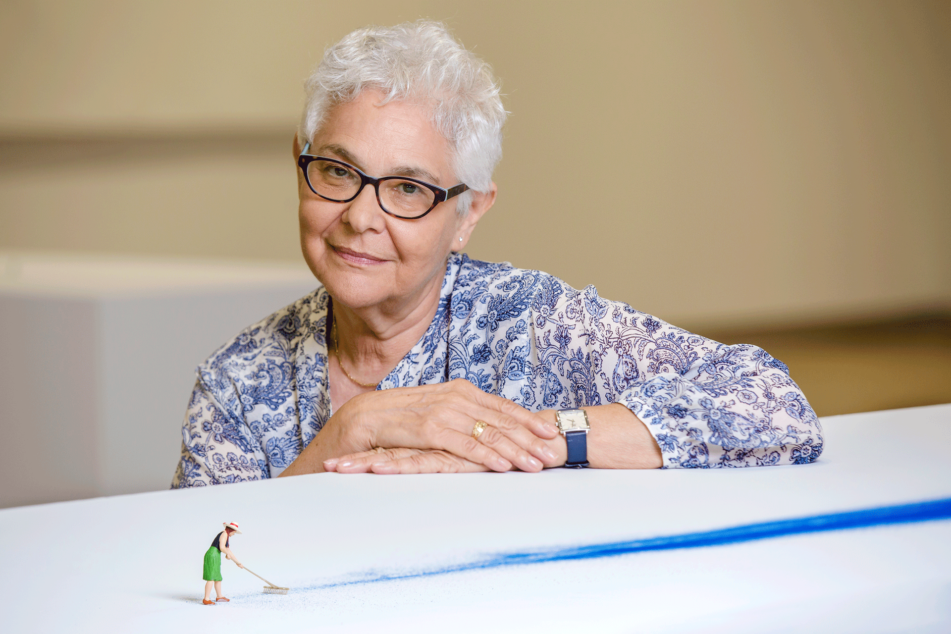 Liliana Porter sits behind her work of a figurine woman sweeping a long trail of blue paint