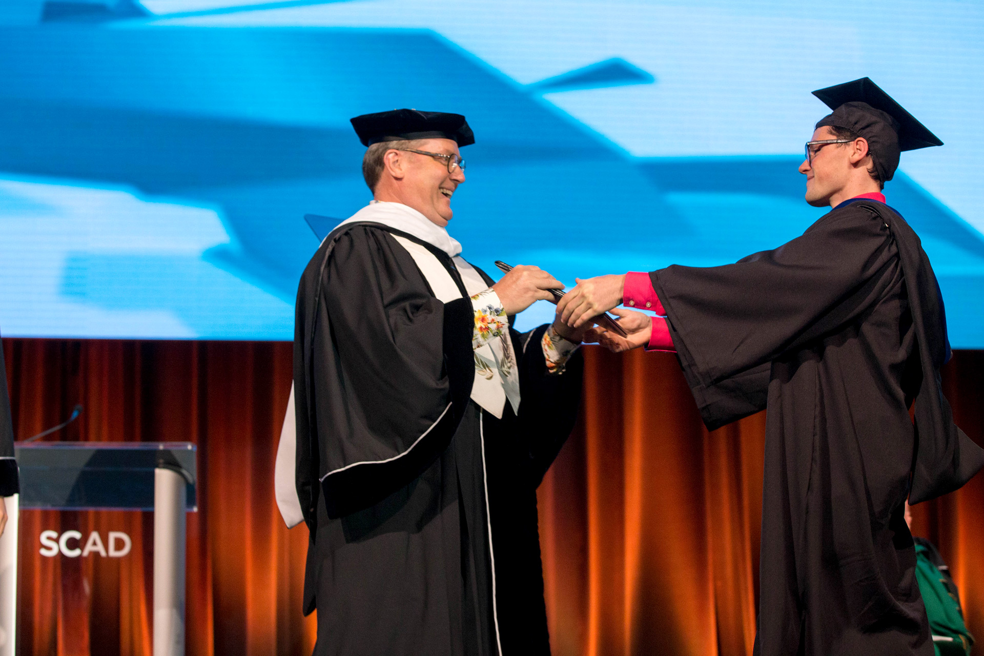 John Lasseter is handed a plaque on stage at SCAD's commencement ceremony
