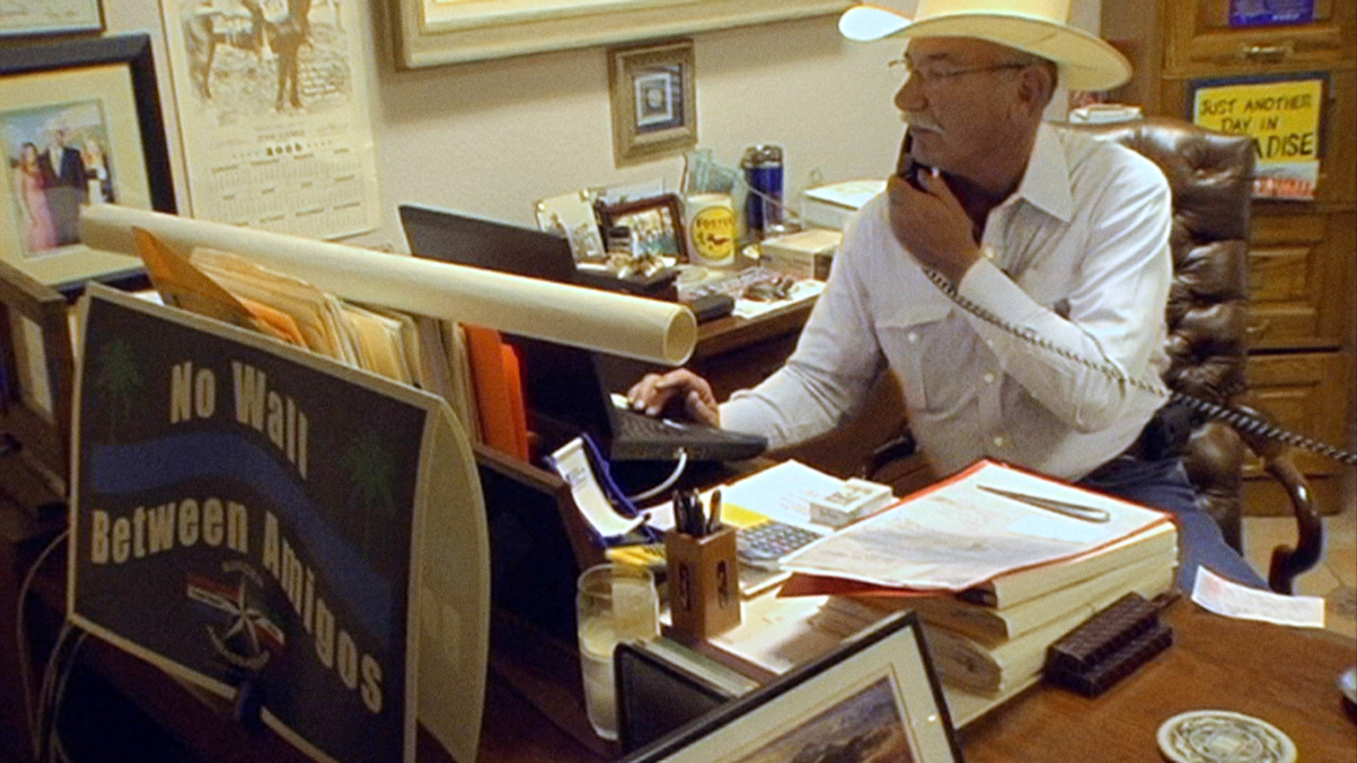 Man in cowboy hat sits at desk while on phone