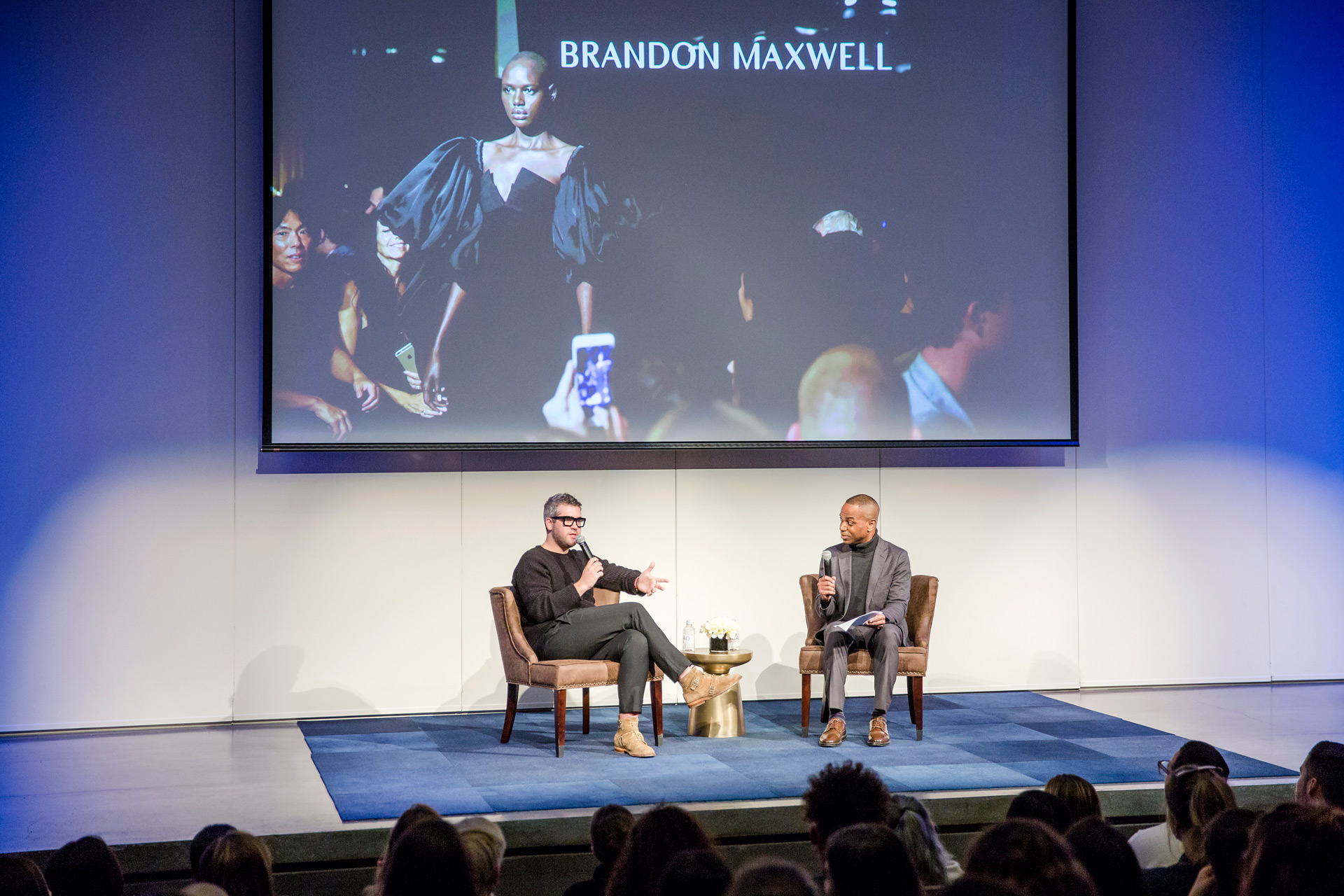 Brandon Maxwell and Ray A. Smith talk on stage, behind them is a large image of a runway show
