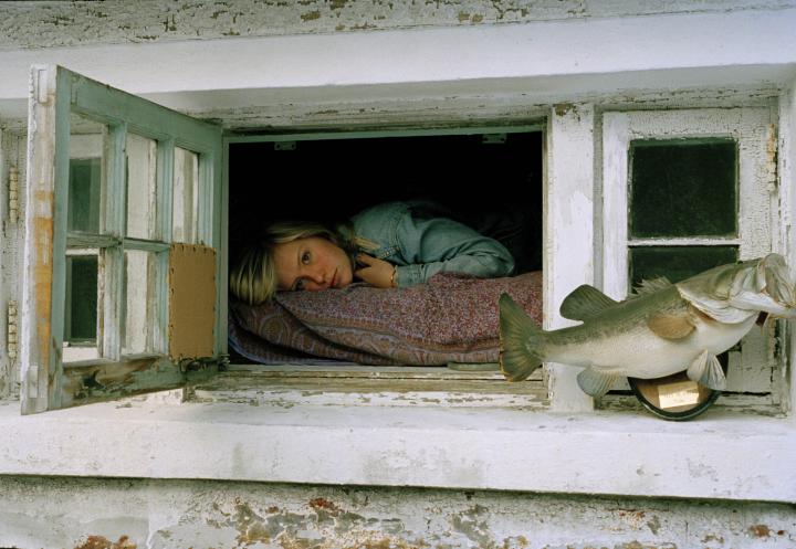 A girl lying on a bed looking out an open window with a mounted bass on the sill