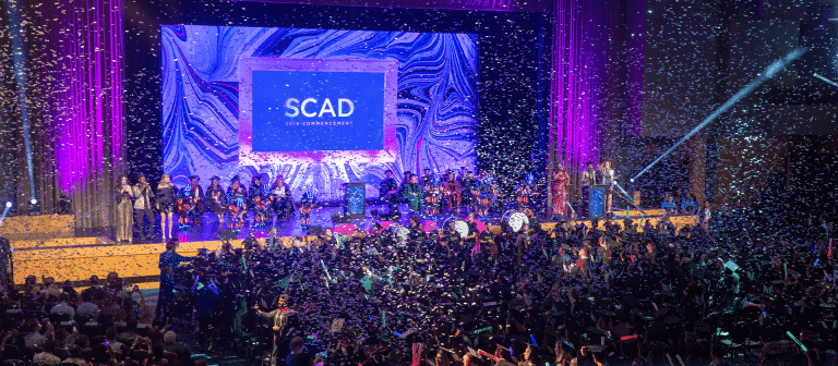 Rows of graduates celebrate with confetti in the air and SCAD banner ahead