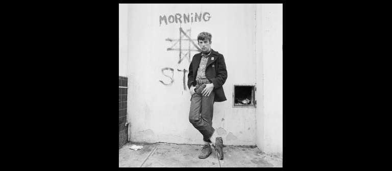 Photographed in black and white, man stands with legs crossed in front of graffitied wall