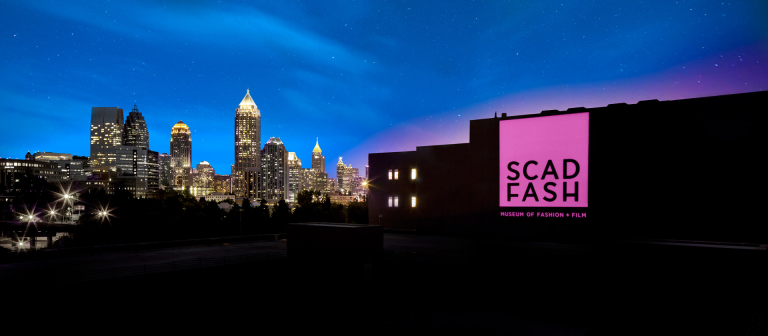 Brightly lit pink SCAD FASH banner on building with Atlanta skyline in background