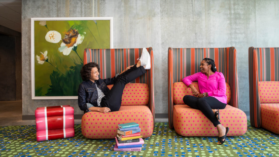Two students sitting on bright furniture in front of a large painting.