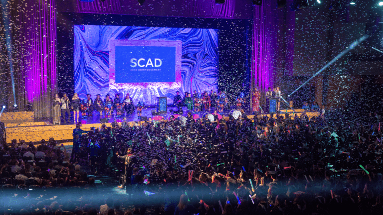 Rows of graduates celebrate with confetti in the air and SCAD banner ahead