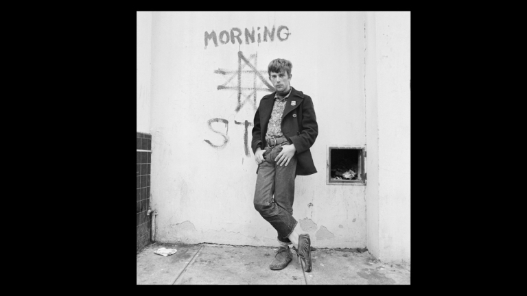 Photographed in black and white, man stands with legs crossed in front of graffitied wall