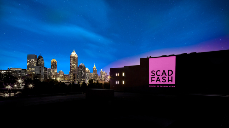 Brightly lit pink SCAD FASH banner on building with Atlanta skyline in background