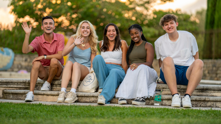 Students sitting on the lawn waving at the camera.