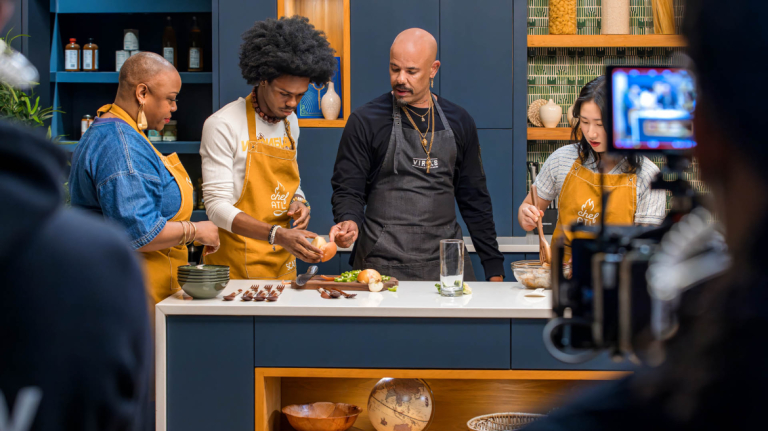 A group of people prepares food on a Chef ATL television cooking set with studio cameras and kitchen equipment.