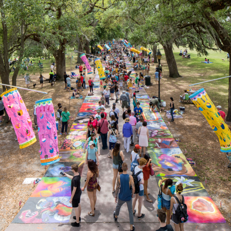 People fill the sidewalk of park during SCAD's sidewalk art festival, pink wind structures flap in the breeze on either side