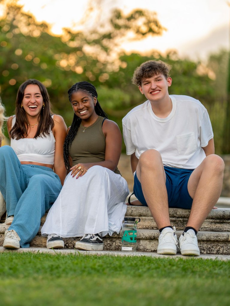 Students sitting on the lawn waving at the camera.